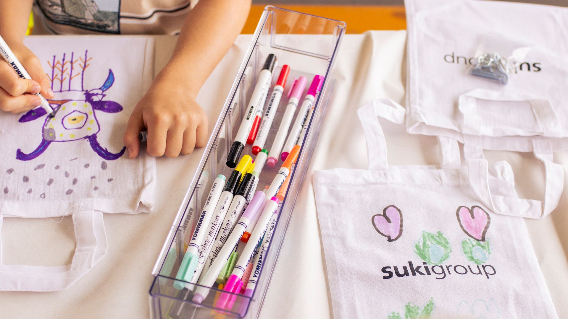 Child coloring a white tote bag with fabric markers, next to a tray of colorful markers and another tote bag with pink hearts and green artwork.