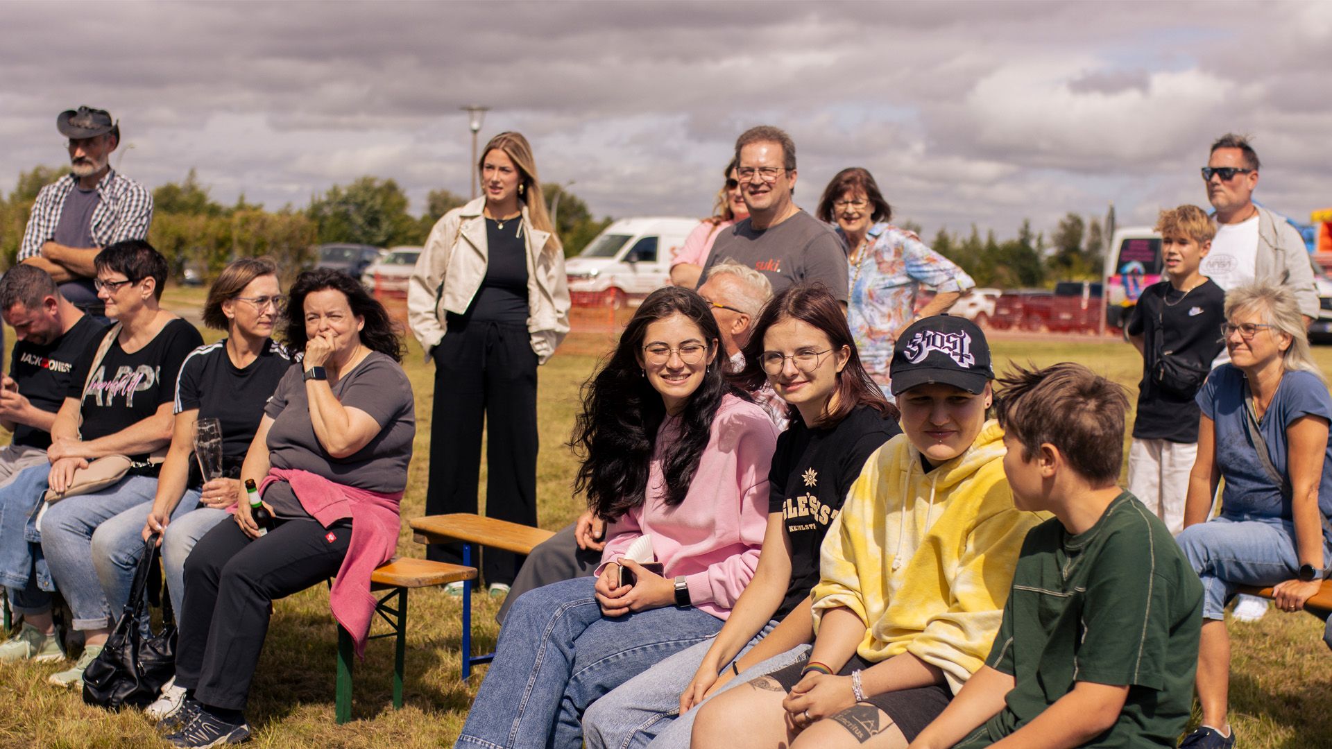 Group of diverse people sitting and standing outdoors on a cloudy day, with some smiling and engaged in conversation.