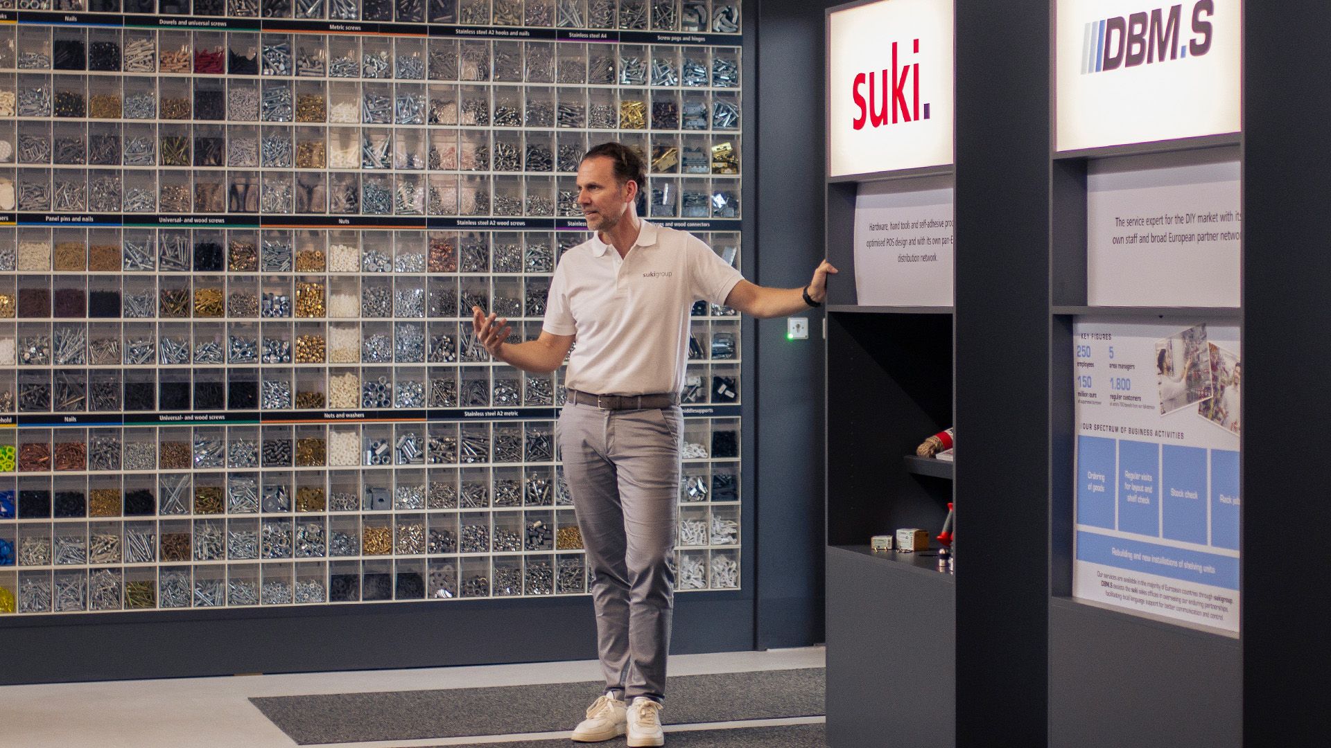 Man in casual attire standing in front of a hardware supply wall with small compartments filled with screws and fasteners.